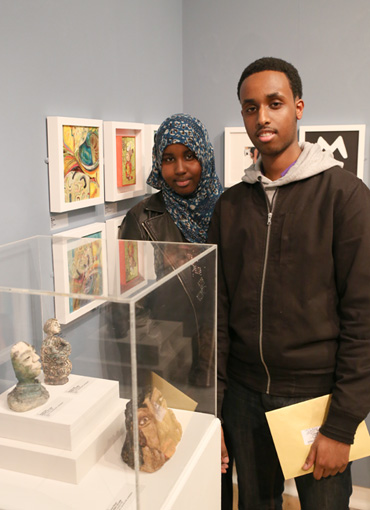 Thumbnail image of Nooridin Abdi (Gateway college student) with his sister, his prize winning 'Ceramic Little Self' is in the cabinet - Little Selves - Preview Photographs